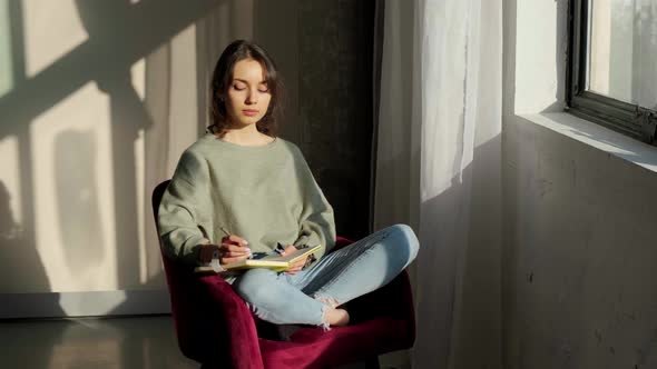 Beautiful Woman Sitting Near Window and Writing Things on Background of Sunset