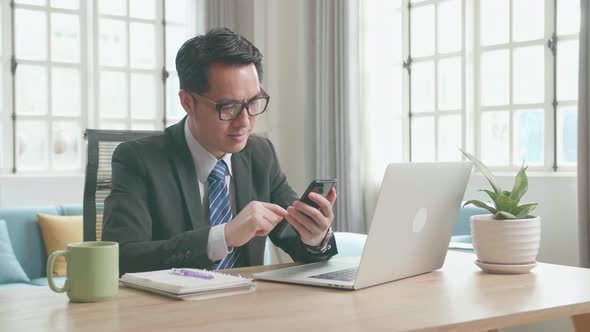 Asian Businessman Using Mobile Phone While Using Computer For Working At Home
