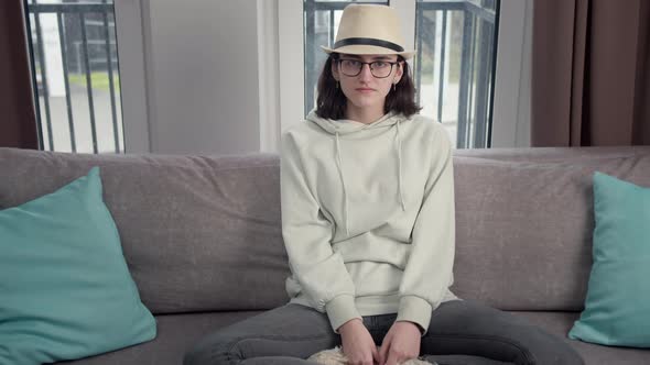Portrait of a Brunette Girl in Glasses and a Hat Sitting on the Couch Looking at the Camera