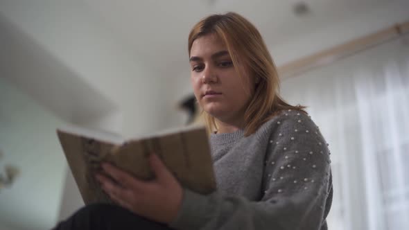 Portrait Plus Size Girl Reading Book Sitting at Sofa in Home. Chubby Woman Holding Book in Hands