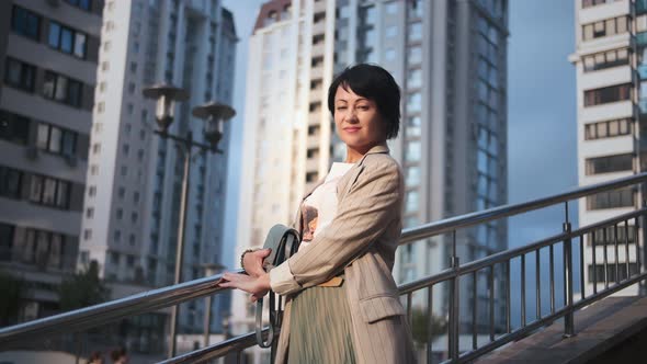 Brunette Stylish Woman Stands in the City Near Houses Looking at the Camera and Smiles Cinematic