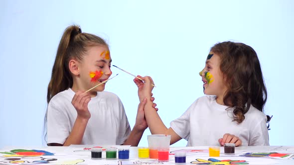 Two Little Girls Sit at the Table, Paint the Tassels on Their Faces. White Background. Slow Motion