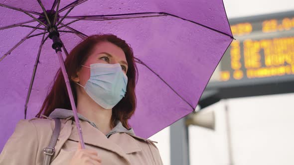 Woman in mask standing at bus stop with umbrella