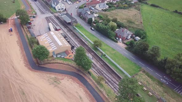 Aerial tracking backward alongside a small train station and tracks, with a busy road behind, and co