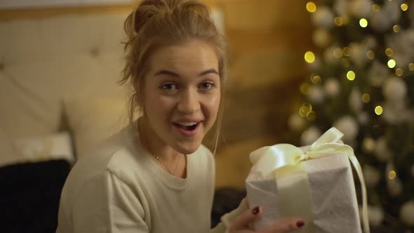 Young Female Shaking New Year Gift Wrapped in White Fabric Expressing Excitement and Joy Being Happy