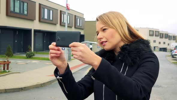 Young Blonde Woman Takes Photos with Phone on Street, Buildings in Background