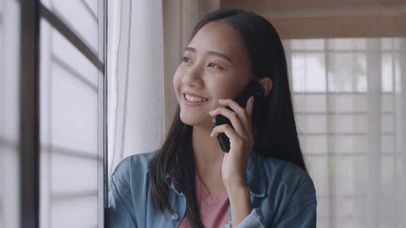 Portrait smiling young Asian woman talking on Phone with friends standing beside a window.