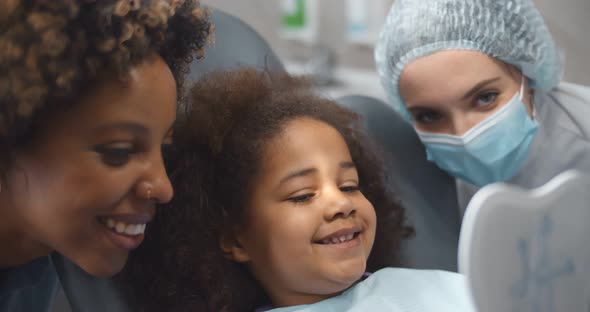 Cute Kid Visiting Dentist with Mom Sitting in Dentist Chair and Looking in Mirror at Beautiful Smile