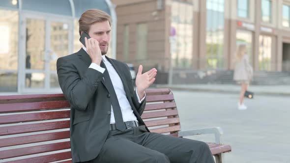 Angry Businessman Talking on Phone While Sitting Outdoor on Bench