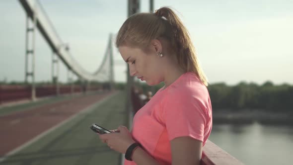 Young Girl in a Sports T-shirt Writes on the Phone While Standing on the Bridge