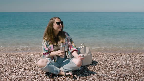Young millennial woman drinking water from a metal bottle and sitting on the seashore on a sunny day