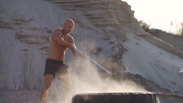 Bald Man Strongman Hits a Hammer on a Huge Wheel in the Sandy Mountains in Slow Motion. Strength and