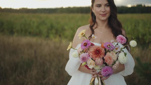 A Beautiful Smiling Bride Stands By the Field and Brings a Delicate Bouquet of Dahlias with Ribbons