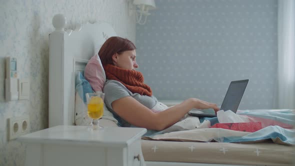 Sick Young Woman Working From Home Laying in Bed with Documents and Laptop. Woman with Warm Scarf on