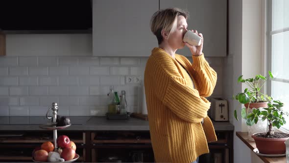 Woman standing at kitchen window, using smartphone, drinking coffee