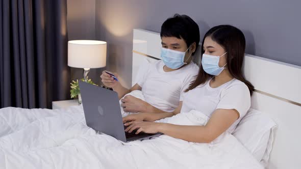 couple with medical masks using laptop to shopping online with credit card on a bed