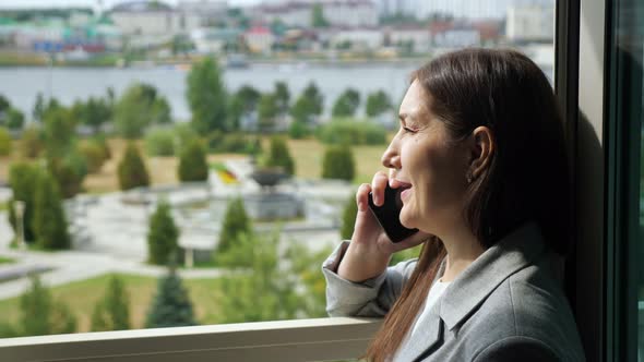 Woman in a Business Suit is Talking on the Phone By the Open Window Against the Background of a