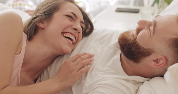 Close Up View of Happy Couple Lying on Bed in Early Morning, Talking Andlaughing. Beautiful Woman