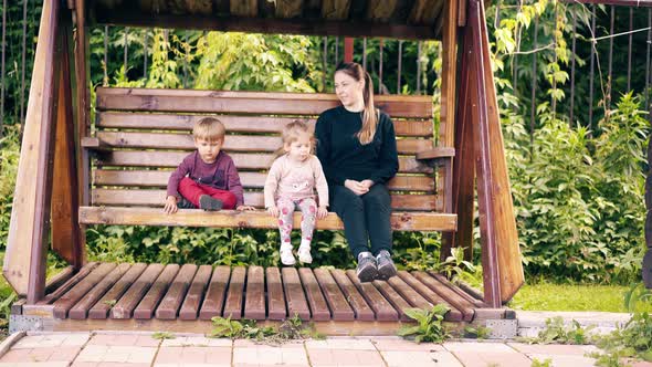 Travelling: Young Pretty Mother with Children, Swinging on a Wooden Swing Near the Summer House