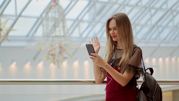 Girl with a Backpack Talking on Video Coll Through Headphones and a Mobile Phone at the Airport