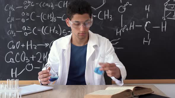 a Male Arab Chemistry Teacher in Glasses Sits at a Table Mixes a Colored Liquid in a Laboratory