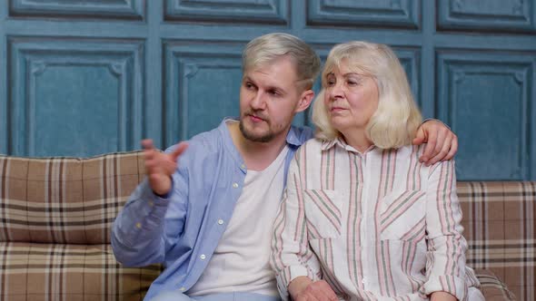 Happy Grown Adult Son Laughing Embracing Talking to Best Friend Older Senior Mother Sit on Sofa