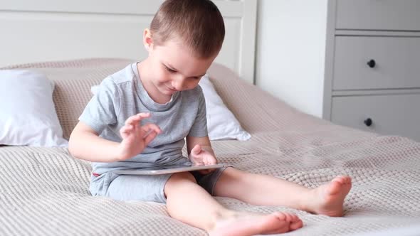 A Small Caucasian Boy in a Light Room in a Gray Tshirt Plays with a Tablet While Sitting on the