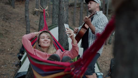 Young Blonde Taking Selfie with Smartphone Camera Swinging in Hammock in Forest While Boyfriend