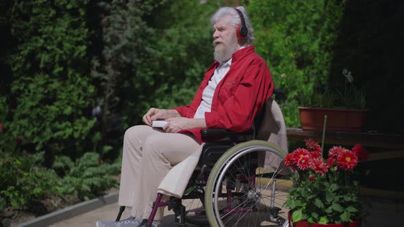 Side View of Positive Confident Impaired Senior Man in Wheelchair Listening to Music in Headphones