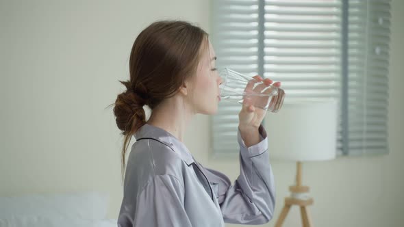 Young Caucasian woman in pajamas drink a glass of water in kitchen.