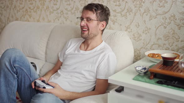 a Young Man with Glasses Sits on the Couch and Plays a Console and Sneezes Many Times