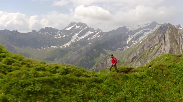 A man in a red jacket running on top of a mountain  towards the camera. In the background you can se