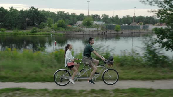 A young couple rides a tandem two-seater bike along the riverbank