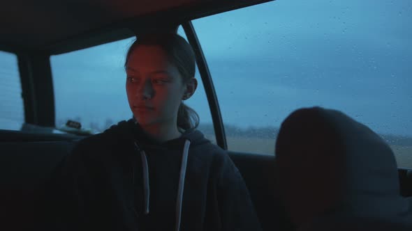 A Cute Young Girl is Sitting in a Car with Raindrops on the Windows and the Reflection of a Fire