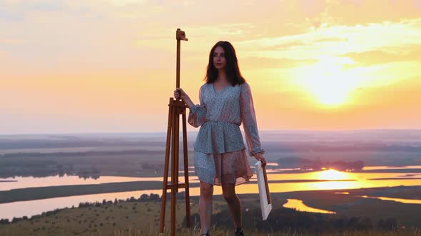 Painting Outside  Young Woman Artist Walks on Sunset Field Holding an Easel and a Canvas