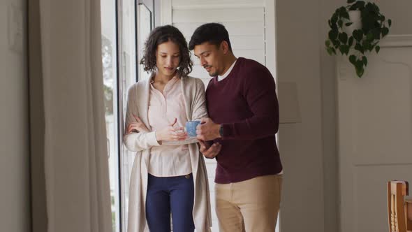 Happy hispanic couple standing in window embracing having coffee