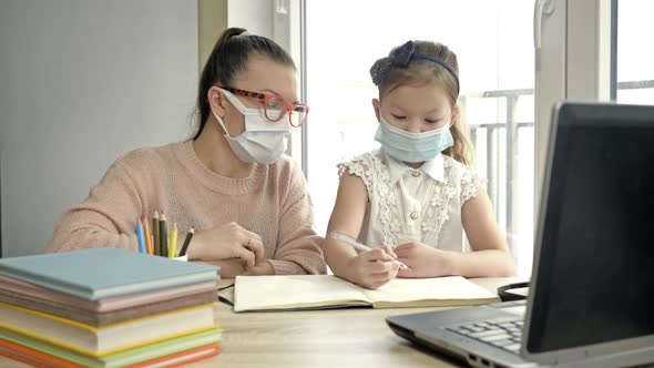 Mom or Teacher or Tutor Helps an Elementary School Student with Her Lessons. Both Have Medical Masks