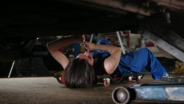 Woman Mechanic with Tattoos Lies Under Car on Car Repair Trolley and Repairs It with Tools