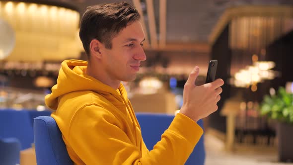 Young Man in a Yellow Hoodie Sits in a Cafe Mall Shopping Center and Uses a Modern Smartphone