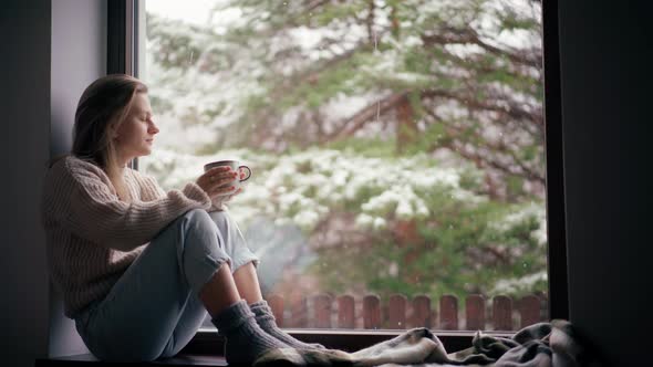 A Beautiful Young Girl Sits on the Windowsill and Drinks Coffee From a Mug