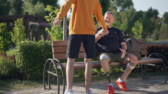 Happy Handsome Mature Amputee Greeting Teenage Boy Sitting on Bench in Sunny Park