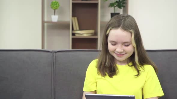 The Little One Smiles and Looks at the Tablet While Sitting on the Couch