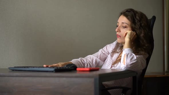 Tired Young Woman with Curly Hair and White Shirt is Working at the Office with Her Computer Sitting