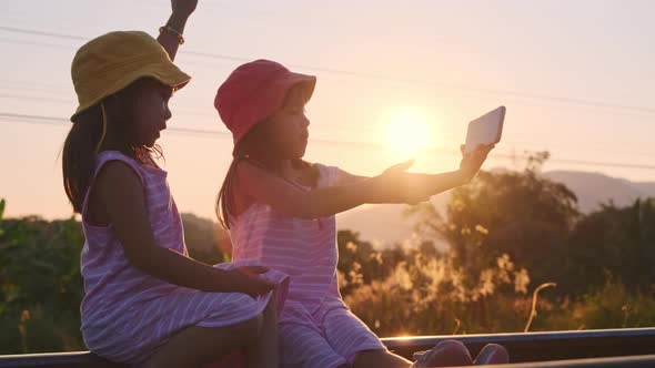 Two sisters sit on the tracks and take selfies together with their smartphones at sunset.