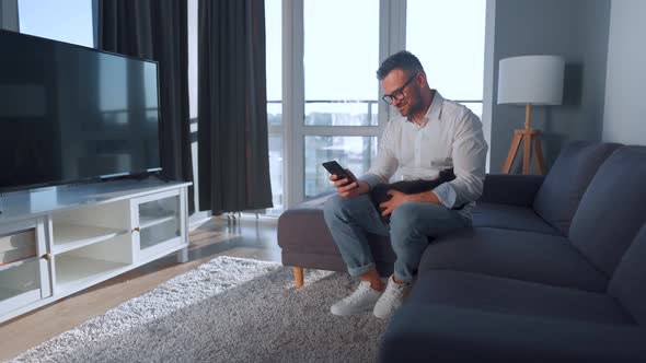 Man with Glasses Sitting on the Couch in a Cozy Room and Using Smartphone for Surfing Internet or