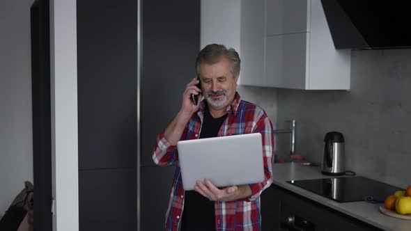 Bearded Old Man Using Laptop in Kitchen and Talking By Phone at the Same Time