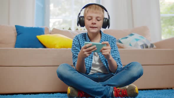 Boy Kid in Headphones Playing Game on Smartphone Comfortably Sitting on Floor