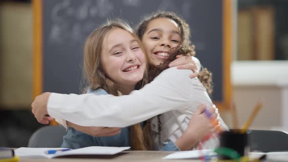 Cheerful Caucasian and African American Schoolgirls Hugging Looking at Camera Sitting at Desk in