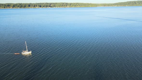 White sailboat towing an orange kayak on calm blue water with distant green trees during the day