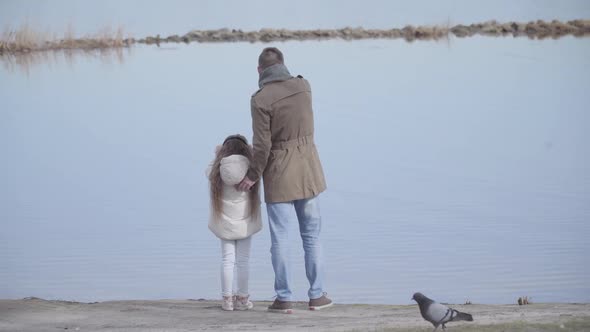 Back View of Young Caucasian Father Standing with Daughter on Riverbank and Talking. Wide Shot of